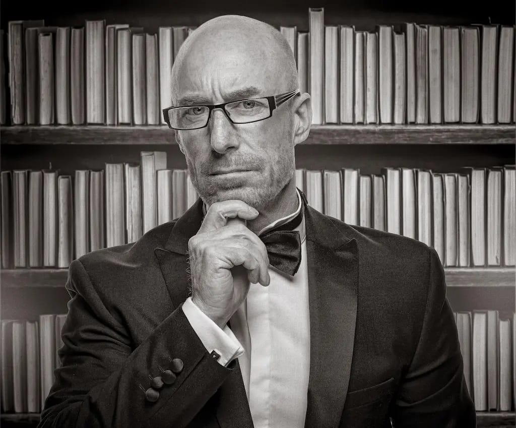 Ritratto in bianco e nero di un uomo calvo con occhiali e papillon, in posa pensierosa con la mano sul mento davanti a una libreria sfocata.