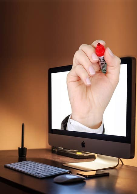 Scrittura digitale: mano con pennarello rosso che emerge dallo schermo di un iMac su scrivania con tastiera e mouse